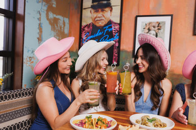 Three women in pink and white cowboy hats cheerfully clinking colorful cocktails over tacos and enchiladas at a lively Mexican-style restaurant