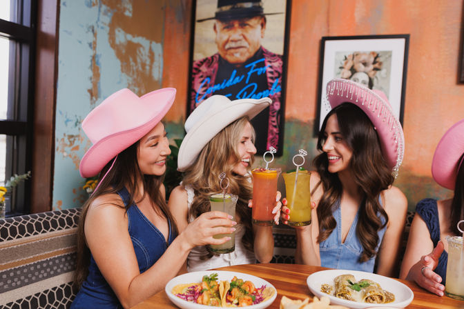 Three women in pink and white cowboy hats cheerfully clinking colorful cocktails over tacos and enchiladas at a lively Mexican-style restaurant