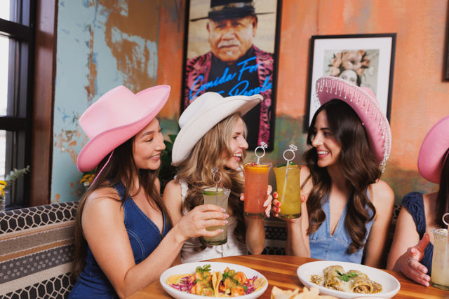 Three women in pink and white cowboy hats cheerfully clinking colorful cocktails over tacos and enchiladas at a lively Mexican-style restaurant