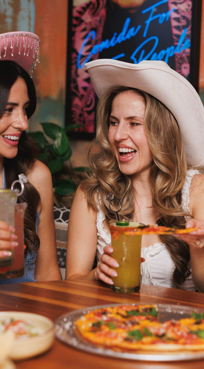 Four friends in pink and white cowgirl hats laughing and toasting cocktails over pizza and tacos at a lively restaurant