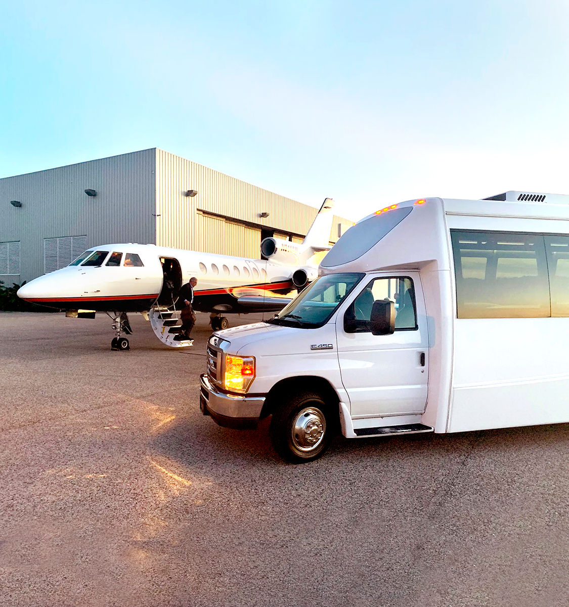 White private jet on an airport tarmac by a hangar at sunset with a passenger descending stairs and a white luxury shuttle van in the foreground