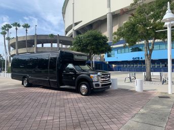 Black luxury shuttle bus parked outside a blue-gated stadium entrance with palm trees and circular ramp under a sunny sky