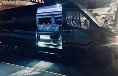 Black luxury shuttle/party van parked at night with sliding door open, glowing blue LED interior and leather bench seating visible in a shopping center parking lot