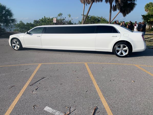 White stretch limousine parked in a sunny beachside parking lot beside palm trees and waterfront, with a small group of people gathered nearby for an event.