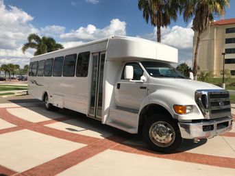 White passenger shuttle bus parked on a sunlit driveway in a palm-tree lined, warm-weather urban setting with a beige hotel building and blue sky