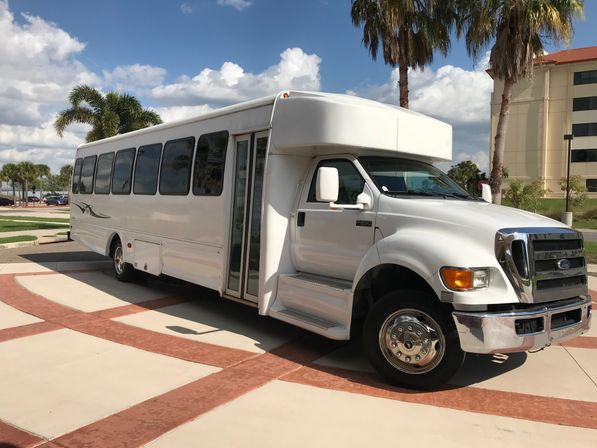 White passenger shuttle bus parked on a sunlit driveway in a palm-tree lined, warm-weather urban setting with a beige hotel building and blue sky