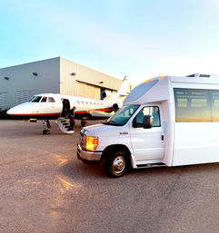 Private jet parked by an airport hangar with a passenger boarding via airstair and a white shuttle van in the foreground on the tarmac at sunset.