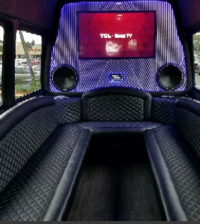 LED-lit luxury party van interior with black quilted leather wraparound seating, central aisle, large flat-screen TV and round speakers, parked in a suburban parking lot visible through side windows.