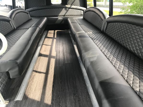 Sleek party bus interior with long black diamond-stitched leather bench seats facing each other, dark wood-look flooring, metal seat tracks, and sunlight streaming through side windows.