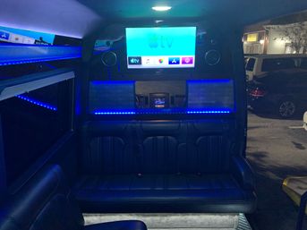 Nighttime luxury van interior with wall-mounted Apple TV, blue LED ambient lighting, built-in speakers and black leather bench seating — party shuttle vibe