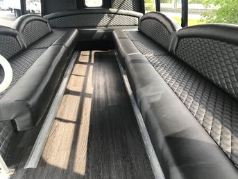 Spacious party-bus interior with black diamond-stitched leather bench seats on both sides, dark wood-look flooring, center aisle and sunlight streaming through windows.