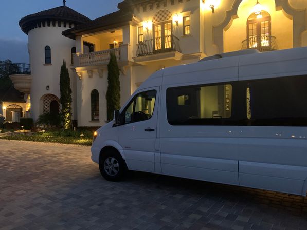 White passenger van parked on a paved driveway in front of a lit Mediterranean-style villa at dusk, showing tiled roof, arched windows, balconies and warm exterior lighting.
