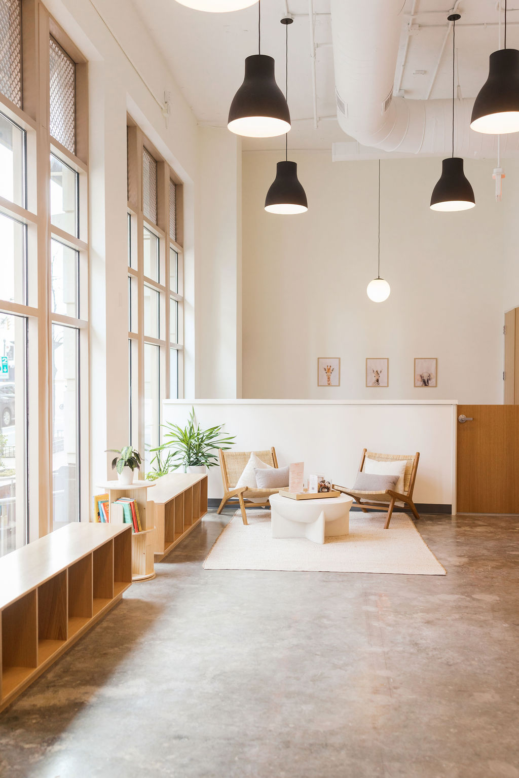 Bright, minimalist Scandinavian-style waiting area with floor-to-ceiling windows, rattan lounge chairs, low white coffee table on a soft rug, indoor plants and black pendant lights over a polished concrete floor.