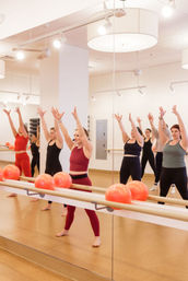 Group barre workout in a bright mirrored fitness studio — instructor in burgundy leads women with arms raised, red exercise balls lined on the barre