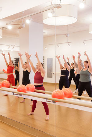 Group barre workout in a bright mirrored fitness studio — instructor in burgundy leads women with arms raised, red exercise balls lined on the barre