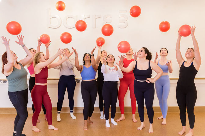 Group of women in colorful activewear in a bright fitness studio tossing red exercise balls overhead, smiling and laughing during a lively group workout.