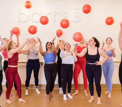 Group of women in colorful activewear in a bright fitness studio tossing red exercise balls overhead, smiling and laughing during a lively group workout.