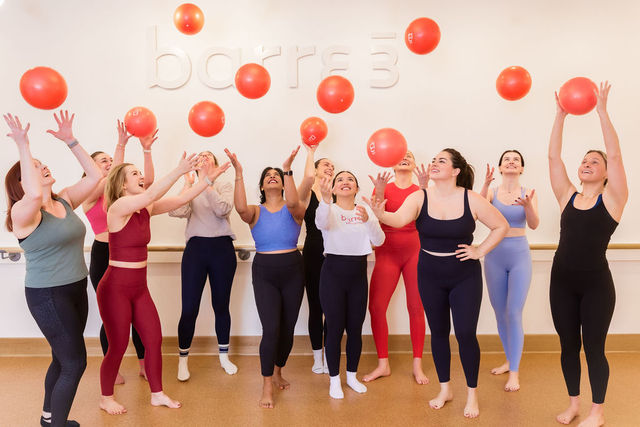Group of women in colorful activewear in a bright fitness studio tossing red exercise balls overhead, smiling and laughing during a lively group workout.