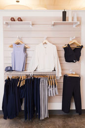 Light-wood retail display of athleisure: lavender tank, cream pullover, black sports bras and leggings, navy and gray workout pants and hoodies on wooden hangers