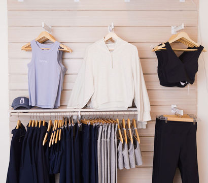Light-wood retail display of athleisure: lavender tank, cream pullover, black sports bras and leggings, navy and gray workout pants and hoodies on wooden hangers