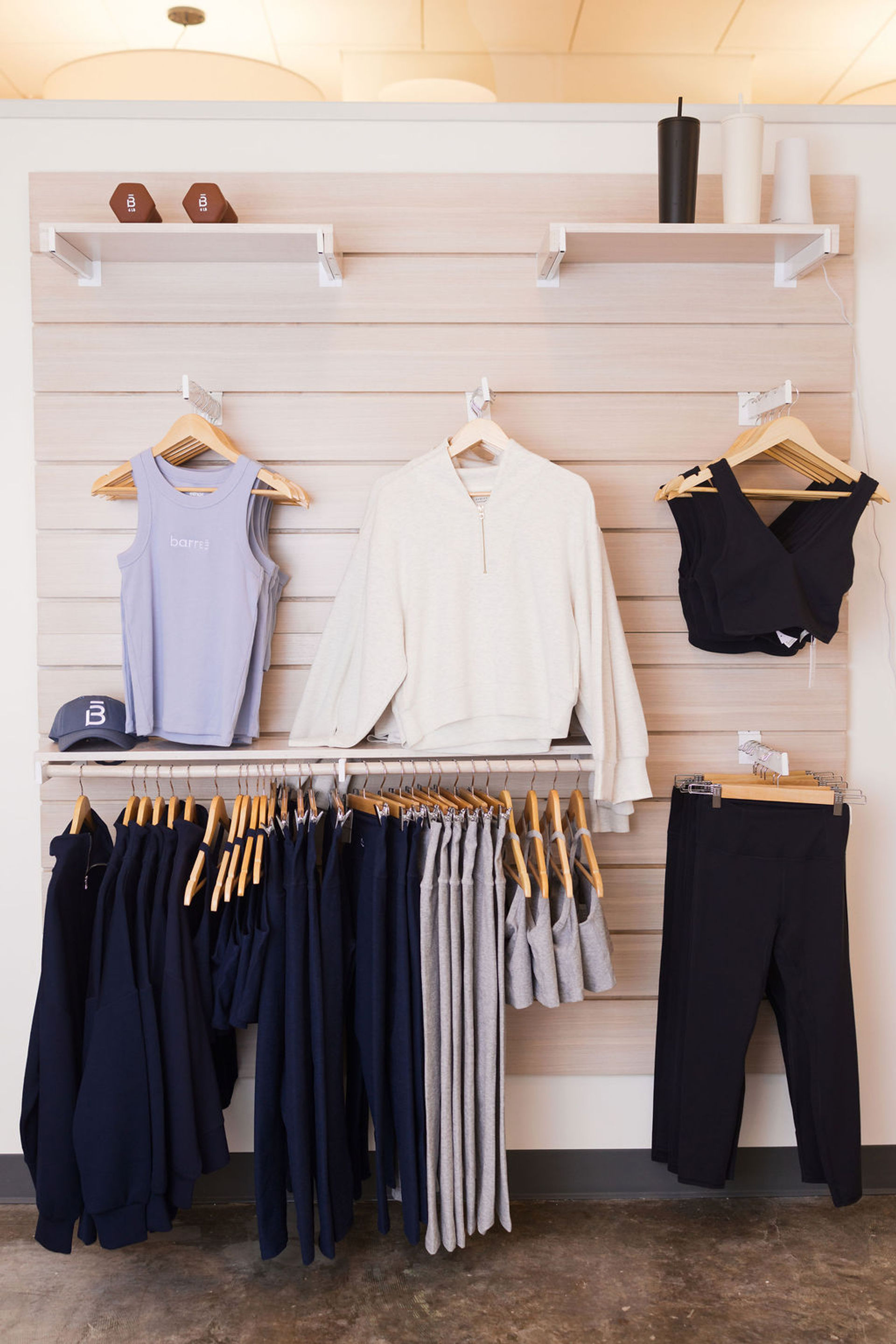 Light-wood retail display of athleisure: lavender tank, cream pullover, black sports bras and leggings, navy and gray workout pants and hoodies on wooden hangers