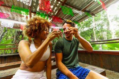 Two friends toasting with shot glasses on a rustic wooden deck at a colorful tropical outdoor bar with red, green and white papel picado decorations and lush jungle foliage.