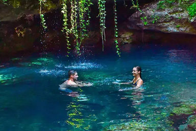Two swimmers enjoying a turquoise cenote in Mexico — clear natural pool under hanging vines and rocky overhang with sunlight reflecting on the water.