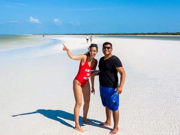 Two people enjoying a wide white‑sand beach under a clear blue sky — a woman in a red swimsuit pointing toward the horizon and smiling, and a man in a black shirt and blue shorts standing beside her with shallow turquoise water and distant shoreline behind them.