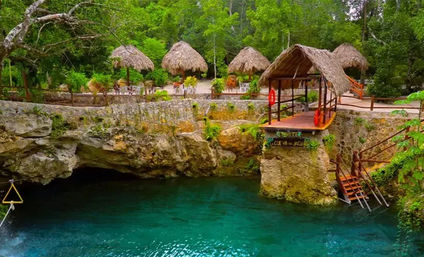 Turquoise cenote surrounded by limestone cliffs and lush jungle, with a thatched-roof wooden dock and ladder and several rustic palapas lining the shore