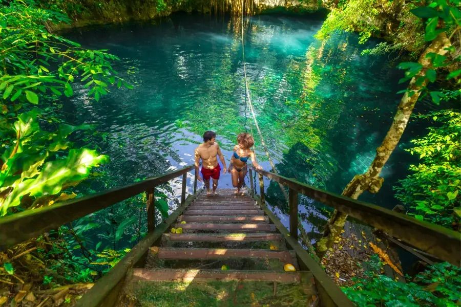 Two swimmers climb wooden steps from a turquoise cenote pool with a hanging rope swing, surrounded by lush tropical jungle and dappled sunlight.
