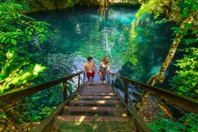 Two swimmers climb wooden steps from a turquoise cenote pool with a hanging rope swing, surrounded by lush tropical jungle and dappled sunlight.