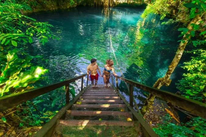 Two swimmers climb wooden steps from a turquoise cenote pool with a hanging rope swing, surrounded by lush tropical jungle and dappled sunlight.