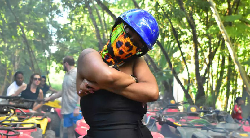 Person in a blue helmet and colorful leopard-print face scarf posing with arms crossed among lined-up ATVs on a sunlit tropical forest trail, outdoor adventure vibe