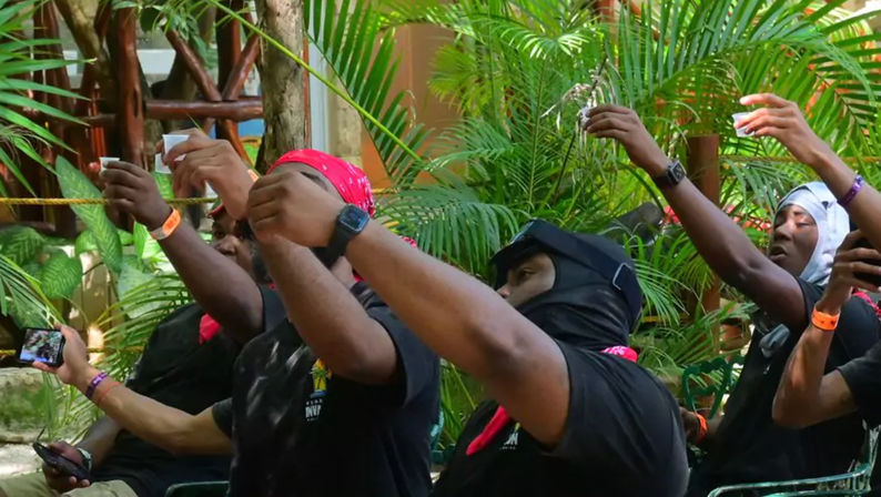Group of people in black shirts and colorful bandanas raising small cups for a celebratory toast at a tropical outdoor bar surrounded by lush palm plants