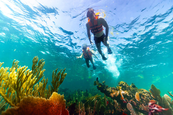 Two snorkelers in orange life vests and fins swimming above a sunlit, colorful coral reef in clear turquoise tropical water