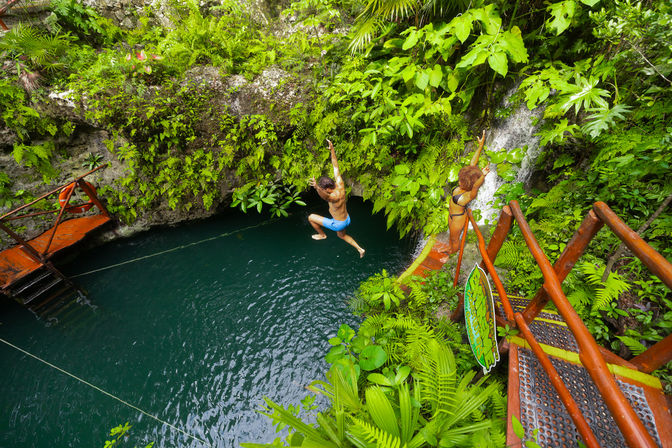 Two people jump from a wooden platform and rope swing into a turquoise tropical cenote fed by a small waterfall, surrounded by lush green ferns and jungle stairs.