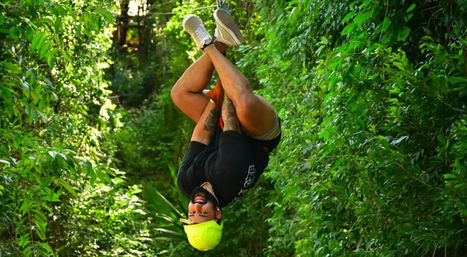 Thrill-seeker hanging upside down on a zipline through a dense tropical rainforest, wearing a yellow helmet and sneakers.
