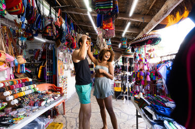 Young tourists laughing as one tries on sunglasses in a colorful outdoor market stall filled with handcrafted bags, beaded bracelets, sombreros and tassel souvenirs