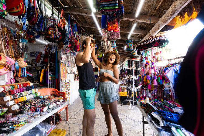 Young tourists laughing as one tries on sunglasses in a colorful outdoor market stall filled with handcrafted bags, beaded bracelets, sombreros and tassel souvenirs