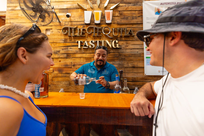Bartender pouring a tequila sample into a plastic cup for two smiling customers at a rustic wooden tequila tasting bar