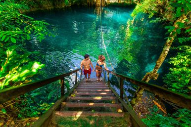 Couple in swimwear climbing wooden stairs into a clear turquoise cenote with a rope swing, surrounded by lush tropical jungle and sunlit emerald water