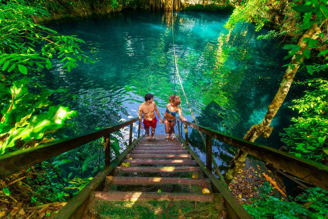 Couple in swimwear climbing wooden stairs into a clear turquoise cenote with a rope swing, surrounded by lush tropical jungle and sunlit emerald water