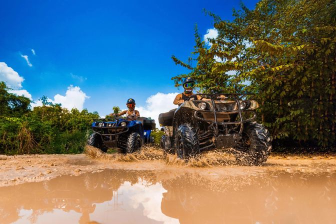 Two riders on ATVs splashing through a large muddy puddle on a tropical off-road trail, dirt flying and bright blue sky overhead.