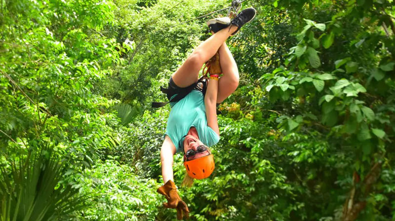 Person ziplining upside down through a lush tropical rainforest canopy, wearing an orange helmet, gloves and sunglasses on a playful adventure