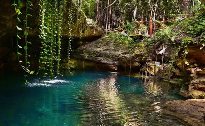 Sunlit turquoise cenote pool in a tropical forest, framed by limestone cliffs and dangling green vines with rippling water reflections.