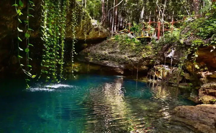 Sunlit turquoise cenote pool in a tropical forest, framed by limestone cliffs and dangling green vines with rippling water reflections.