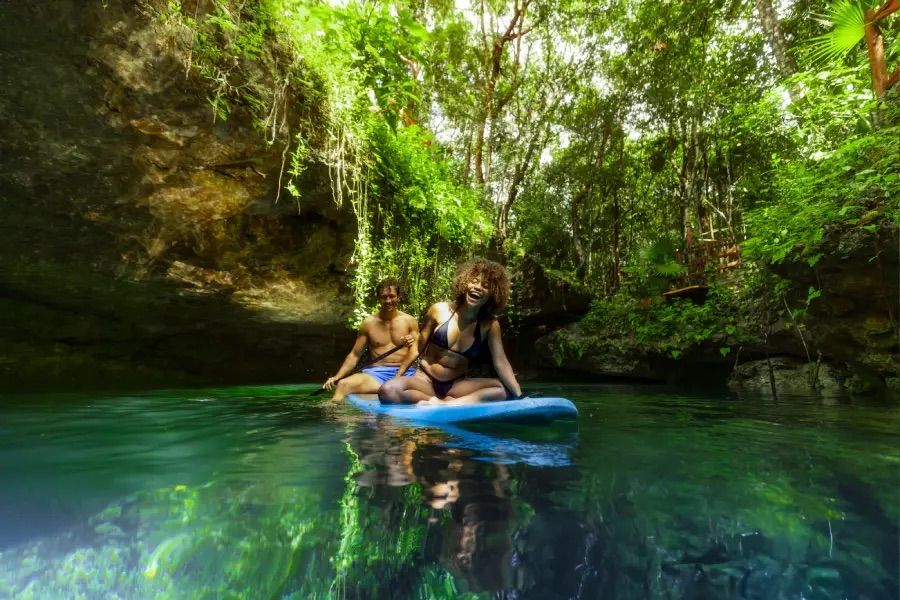 Two people laughing on a blue paddleboard in a crystal-clear tropical cenote, surrounded by lush green jungle, hanging vines and rocky overhangs — paddleboarding in a serene jungle lagoon.