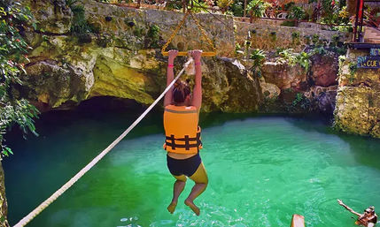 Person in an orange life vest swinging on a rope over bright turquoise cenote water inside a limestone grotto rimmed with tropical plants