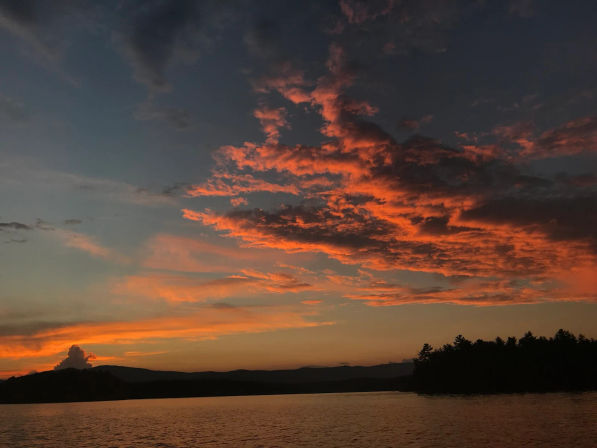 Fiery orange sunset over a calm lake, dramatic glowing clouds streaking the evening sky above a dark silhouetted treeline and distant hills.