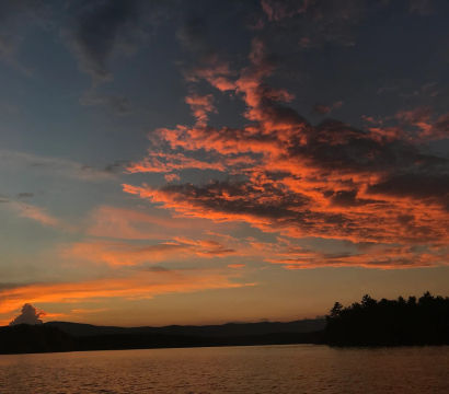 Fiery orange sunset over a calm lake, dramatic glowing clouds streaking the evening sky above a dark silhouetted treeline and distant hills.